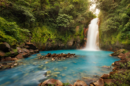 Wasserfall m&uuml;ndet in einem blauschimmernden See, umgeben von gr&uuml;nem dichten Wald
